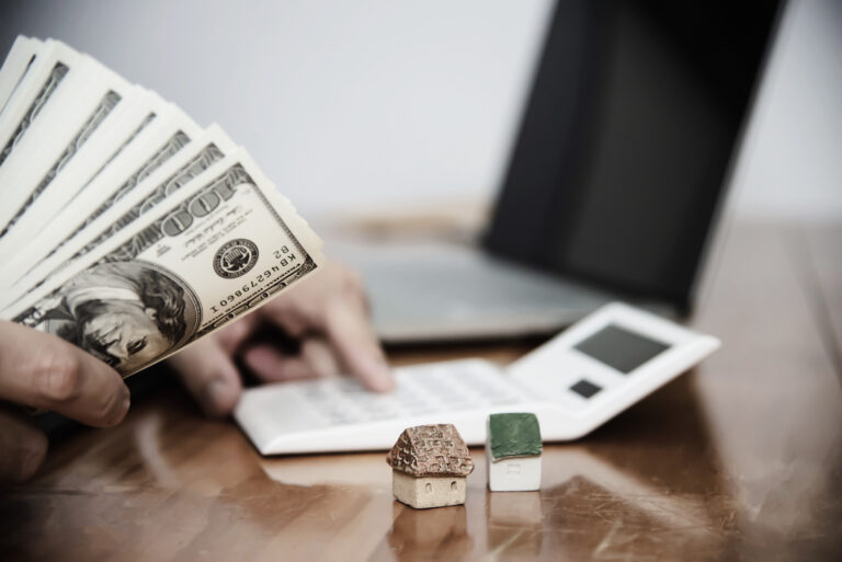 A person holds a stack of dollar bills while calculating finances next to miniature house models, representing economic considerations—linked to "How the 2024 Elections Could Impact Financial Advisors"